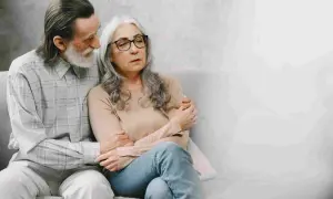 Coppia anziana seduta sul divano. Lui la conforta, forse parlando di come la pensione di invalidità è reversibile.