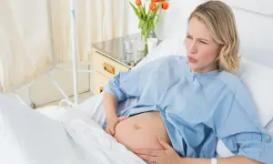 Kobieta w szpitalnym łóżku, z bólem na twarzy, trzymająca się za brzuch. Zastanawia się, jak wywołać poród.