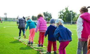 Niños corriendo en una escalera de agilidad en un campo de césped artificial, ideal para aprender inglés en lugares divertidos.