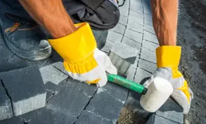 Pracownik w żółtych rękawicach układa kostkę brukową na betonie, używając gumowego młotka.