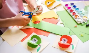 Manos pequeñas cortando papel para manualidades para niños de 3 a 5 años. Pinturas, tijeras y frutas de papel decoran la mesa.