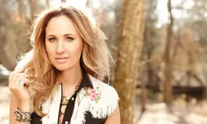 Jessi Alexander, a blonde woman with striking eyes, poses outdoors in a fringed vest adorned with floral embroidery.