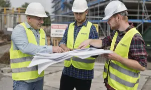 Trzech budowlańców w kaskach i kamizelkach omawia plany. Jeden z nich wskazuje palcem na dokumenty, ustalając szczegóły projektu i prowizję.