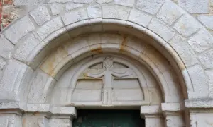 Kamienna płaskorzeźba postaci z koroną na głowie, rozłożonymi ramionami i ozdobnym pasem, osadzona w łuku portalu. To przykład sztuki romańskiej.