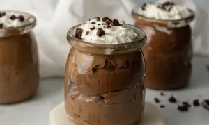 Czekoladowy pudding proteinowy w słoiku, udekorowany bitą śmietaną i kawałkami czekolady.