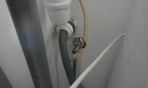 Szara rura odpływowa pralki podłączona do syfonu pod zlewem. Widoczny żółty przewód i biały kabel.