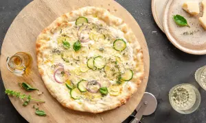 Pizza z cukinią, cebulą i cytryną na kremowym, białym sosie do pizzy. Obok buteleczka oliwy i kawałki parmezanu.
