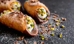 Trois cannoli salés, garniture crémeuse, pistaches concassées et copeaux de chocolat, saupoudrés de sucre glace sur fond sombre.