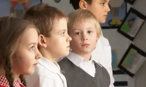Cztery dzieci w szkolnych strojach, skupione na czymś poza kadrem. W tle widać prace plastyczne, sugerujące naukę i nauczanie indywidualne.