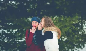 Dwie kobiety szepczą sobie do ucha na tle drzew.