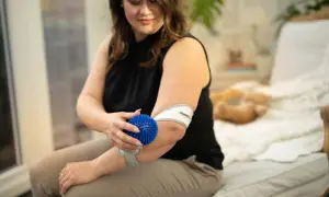 Frau massiert ihren Unterarm mit einem blauen Igelball und trägt eine Bandage.