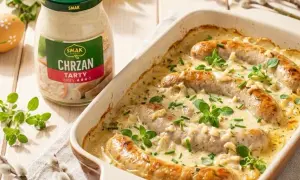 Biała kiełbasa w sosie chrzanowym, zapiekana w naczyniu, z dodatkiem ziół. Obok słoik sosu.
