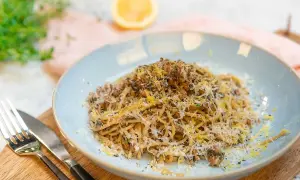 En tallrik med saffranspasta vegetarisk, toppad med riven ost och citronzest. Serveras med bestick på en träskärbräda.