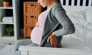 Femme enceinte assise sur un lit, tenant son ventre et se tenant le bas du dos, souffrant peut-être d'hémorroïdes.