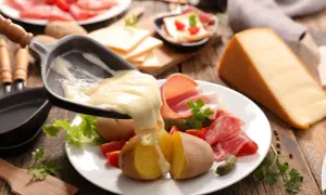 Roztopiony ser raclette spływa na ziemniaki i wędliny. Obok kawałek sera i talerz z dodatkami.