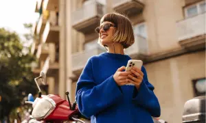 Frau mit blondem Bob, Sonnenbrille und blauem Pullover, die auf ihrem Handy scrollt. Ein rotes Moped steht daneben. Ein toller Look, wie man einen blauen Pullover kombinieren kann, Herren-Style inspiriert.