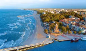 Widok z lotu ptaka na **latarnię morską Kołobrzeg** i wybrzeże Bałtyku. Widać plażę, molo i miasto.