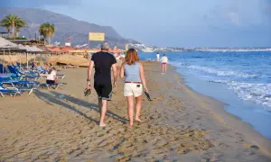 Spacerują plażą, zastanawiając się, ile wynoszą wczasy pod gruszą. W tle widać morze, góry i kurort.
