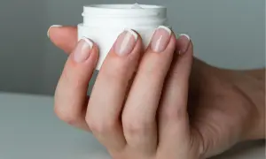 Dłoń z paznokciami w stylu francuskim trzyma słoiczek z kremem. Dowiedz się, jak nakładać bazę kauczukową dla idealnego manicure.
