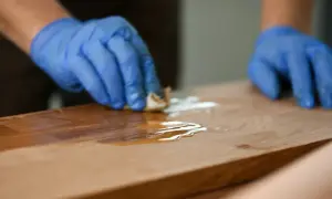 Ręce w niebieskich rękawiczkach nakładają olej na drewniany, porysowany blat kuchenny, przywracając mu blask.