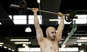 Uomo calvo con barba solleva bilanciere sopra la testa, concentrato sull'allenamento forza esplosiva.