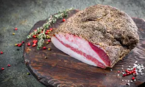 Pancetta co to? Wędzony kawałek boczku z widocznymi warstwami tłuszczu i mięsa, przyprawiony pieprzem, leży na drewnianej desce z ziołami i ziarnami pieprzu.