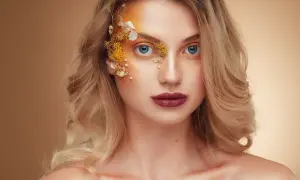 Fotografia beauty: kobieta z niebieskimi oczami, ozdobiona kwiatami i złotym makijażem na twarzy.