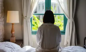Kobieta w szlafroku siedzi na łóżku, patrząc przez okno. Może to być obraz odzwierciedlający zaburzenia psychogenne objawy, samotność i refleksję.