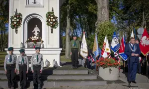 Uroczystość przy kapliczce. Czy imię Bruno jest święte? Dzieci w harcerskich mundurkach i dorosły z łańcuchem na szyi stoją przed kapliczką z figurą i flagami.