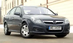 Ciemnoszary Opel Vectra C z polskimi tablicami WE 3439F. Widać jego przednie reflektory, atrapę chłodnicy z logo Opla i zderzak.