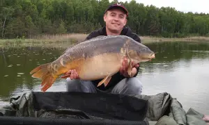 Szczęśliwy wędkarz prezentuje okazałego karpia złowionego na łowisku tuszynek. W tle jezioro i las.