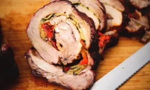 Coppa di maiale allo spiedo, succulenta e ripiena di verdure, pronta per essere affettata su un tagliere di legno.