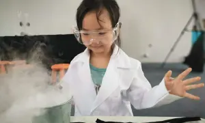 Niña con bata y gafas realiza experimentos para niños de 7 años, creando humo blanco de un cubo.