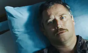 Mężczyzna z wąsami leży na niebieskiej poduszce, jego oczy patrzą w górę. Może to scena z filmu "Dzień świra", gdzie reżyser ukazuje codzienne zmagania bohatera.