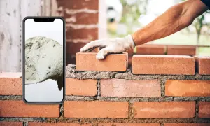Teléfono móvil muestra cemento, mano con guante coloca ladrillo en pared.