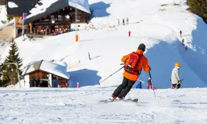 Narciarz w pomarańczowej kurtce z plecakiem zjeżdża po ośnieżonych stokach narciarskich w Austrii. W tle widać górskie chaty i innych narciarzy.