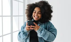 Uśmiechnięta kobieta z burzą loków przegląda telefon, szukając inspiracji, jak udostępnić czyjąś relację na Instagramie.