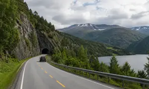Svensk bil på väg mot en tunnel i Norge. Vacker bergig natur med sjö och moln.