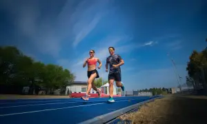 Coppia corre su pista blu, un allenamento intenso per la loro prossima corsa a tappe. Cielo azzurro e nuvole bianche.