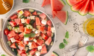 Erfrischender Wassermelonen Feta Salat mit Gurkenstücken und Minze, garniert mit Pfeffer. Daneben ein Glas Dressing und Honig.