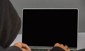 Osoba w kapturze pisze na laptopie z czarnym ekranem. Ciemność skrywa jej działania.