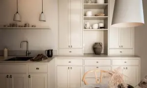 Ett kök med shakerinredning i ljusa toner. Öppna hyllor med keramik, en diskho med kromad kran och en stor taklampa.