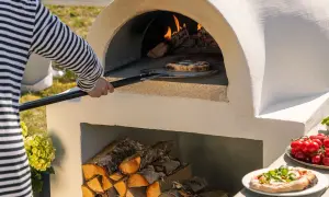En person med randig tröja använder en spade för att lägga in en pizza i en nybyggd pizzaugn. Ved ligger staplad under ugnen.