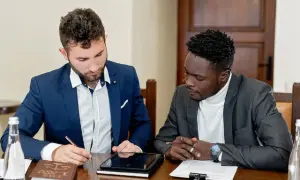 Dwóch mężczyzn w garniturach przy stole, jeden pisze, drugi patrzy. Rozmawiają o tym, jak zwracać się do notariusza.