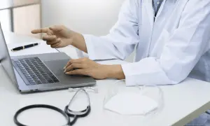 Lekarz w białym kitlu przy laptopie wyjaśnia, czy do poradni dermatologicznej potrzebne jest skierowanie. Na biurku stetoskop i maseczka.