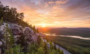 Solnedgång över en skogklädd dal med en sjö. Klippor och träd i förgrunden. En vacker vy som ger fakta om Finland.