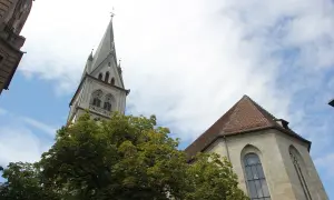 Der Turm der Stephanskirche Konstanz ragt mit seiner Spitze in den blauen Himmel. Ein Baum mit dichtem Laub steht davor.