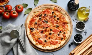 Pyszna pizza bez glutenu z pomidorami i oliwkami, gotowa do pokrojenia. Obok świeże pomidory, oliwa i grissini.