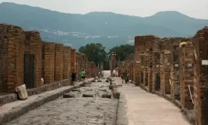Besucher spazieren auf der antiken, kopfsteingepflasterten Straße von Pompeji heute, umgeben von den Ruinen römischer Gebäude. Berge im Hintergrund.
