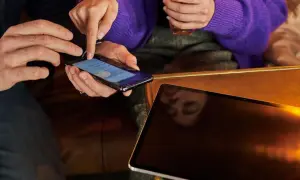 Dwie osoby przeglądają ekran telefonu. Obok leży tablet, na którym widać odbicie twarzy. To ciekawy screenshot.